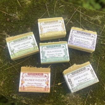 Assorted Old Factory goat milk soap boxes placed on a mossy rock outdoors.