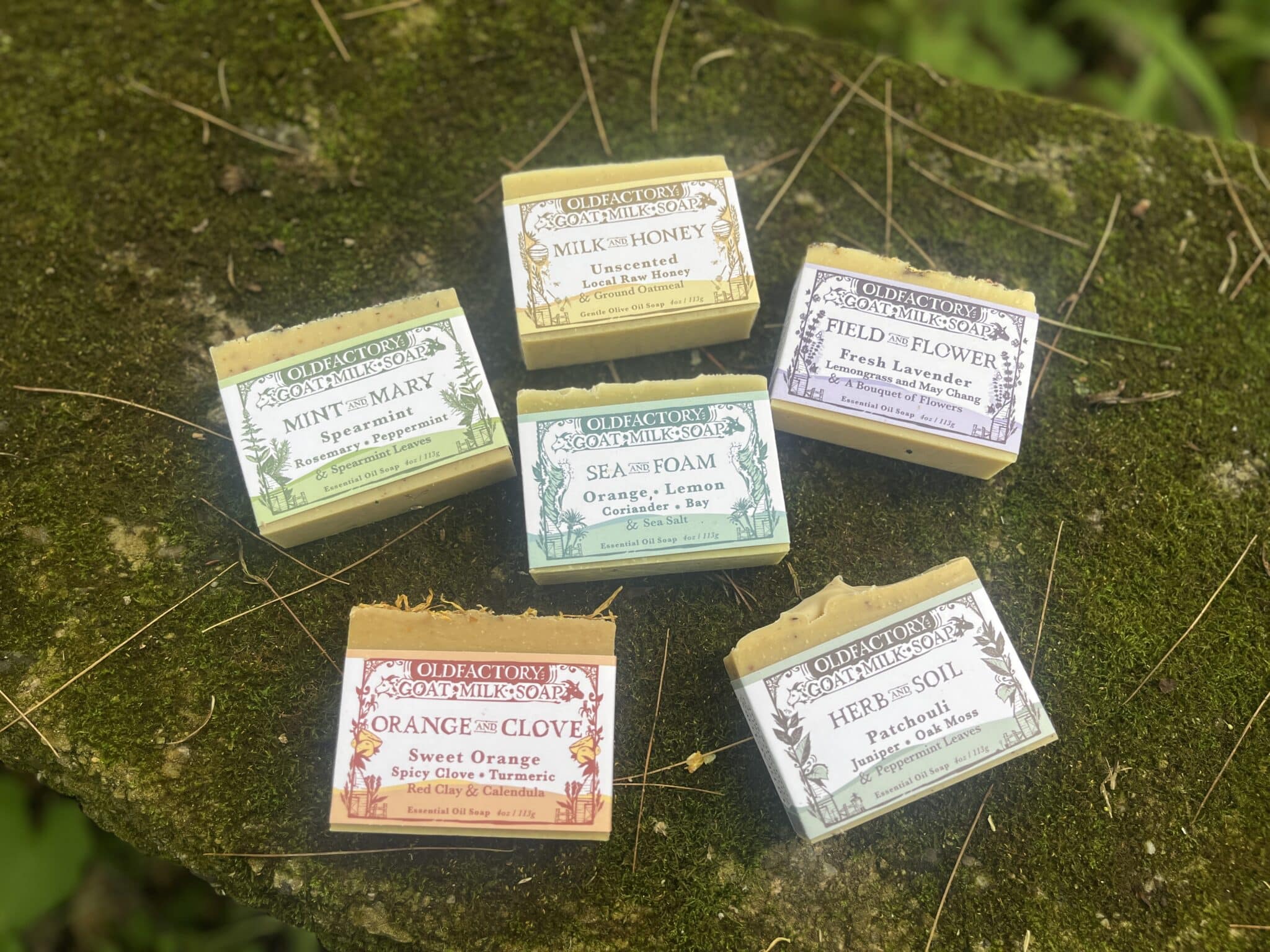 Assorted Old Factory goat milk soap boxes placed on a mossy rock outdoors.